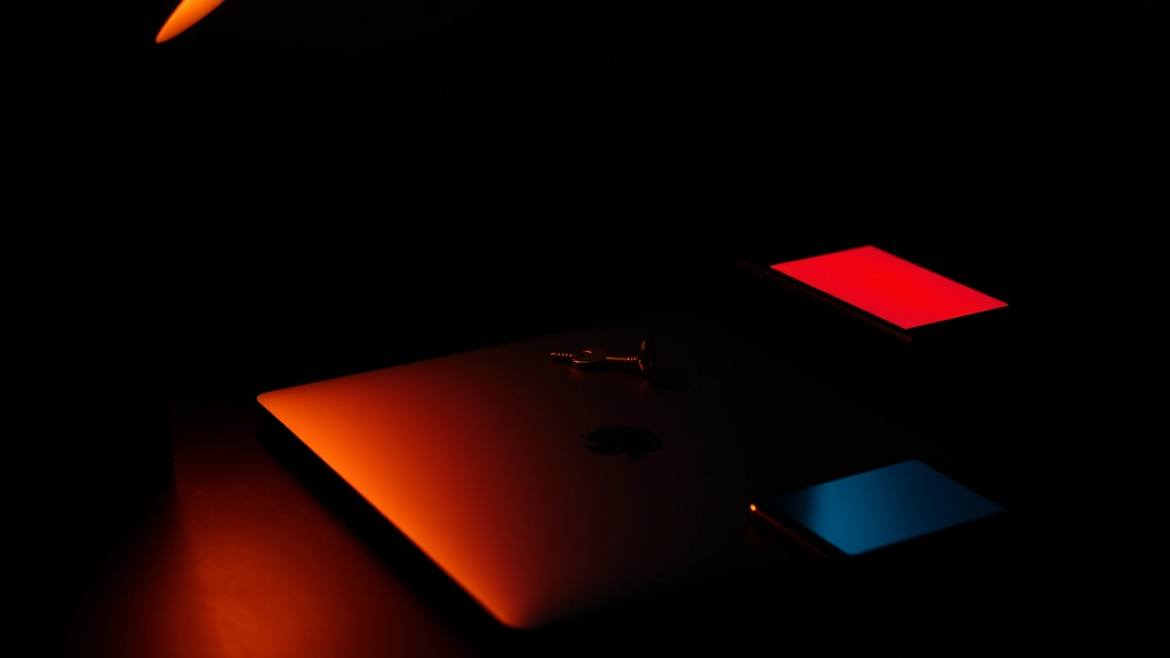 A dark desk lit from the left holding a closed laptop, a small brass key lying on the lid and a phone face down beside it, a faint crimson glow from a screen somewhere off frame